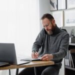 man on the phone while writing on his notebook_Pexels